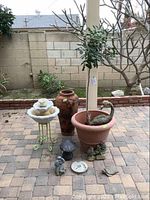 Overview of yard decor lot showing large terracotta pot, white two-tier fountain on green metal stand, large planter with small olive tree, elephant sprinkler, mushroom and frog figurines, and garden stones.
