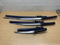 Three Japanese-style swords laid side by side on a light wooden surface. Each sword has a blue scabbard and a black handle, visible inscriptions on the blades.