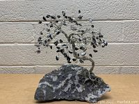 Front angle showing the rock tree sculpture with twisted wire branches and black stone leaves on a textured gray and white stone base.
