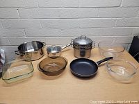 Photo showing various pots, pans, and glass bakeware arranged on a wooden surface against a brick background