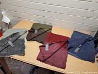 Four new men's ultra soft knit scarves in black/white, olive green, burgundy, and navy blue, all with tags attached, displayed on a table.