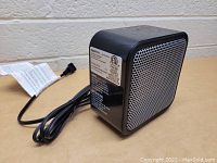 Front and side view of small cube-shaped desktop heater with black body and silver grille, power cord coiled beside it.