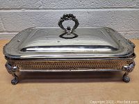Top down view of the vintage silver plated serving set with glass dish, showing the lid and ornate handle on silver plated stand with legs and handles.