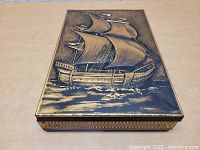 Front top view of rectangular metal trinket box showing embossed sailing ship on lid with gold-tone accents.
