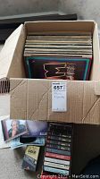 Box containing vertically stacked vinyl 33 RPM albums with varied cover art and a small pile of assorted audio cassettes beside the box.