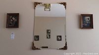 Rectangular wall mirror with bevelled edge and four ornate moulded corners in antiqued gold finish. Two small framed prints visible on either side of the mirror on the wall.