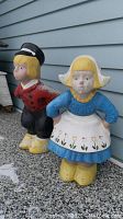 Full view of Dutch boy and girl cement garden statues together outside on concrete surface next to blue house siding.