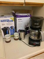 Photo showing the Proctor Silex cordless electric kettle in its original box, the Rival coffee maker box, the Black and Decker coffee maker, a stainless steel travel mug, and two ceramic mushroom coffee cups arranged on a kitchen counter.