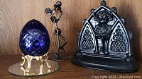 Blue cut to clear glass egg on ornate metal legs, metal nuts and bolts figurine, and painted cast metal Lincoln Imp doorstop on wooden surface