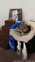 Photo showing brown foam dog steps with fabric cover, cream fabric covered dog bed holding cleaning products and blue dog coat, with framed picture of a dog in background.