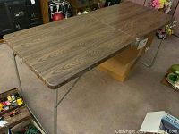 Full view of mid century folding table with wood grain veneer top and metal folding legs intact, set up on carpeted floor.