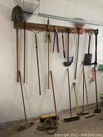 Wide view showing various garden tools hanging on a wooden rack, including rakes, shovels, brooms, and hand tools.