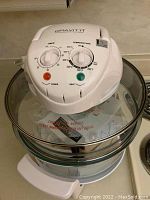White Gravitti halogen oven lid with timer and temperature control dials on top of clear glass bowl.