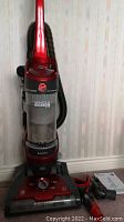 Front view of upright Hoover vacuum cleaner showing red body, hose coiled on side, clear dirt canister, control knob, and attachments placed beside it, along with the manual.