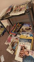 Books and magazines of knitting patterns stacked on and under a small plastic shelf. The covers show various knitting designs and styles. Carpet and part of a rug visible on the floor.