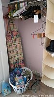 Fabric hanging storage organizer with six compartments with a black top hat on one shelf, an ironing board with floral and plaid cover, a plastic laundry basket with various laundry items, and assorted hangers on a metal rod.