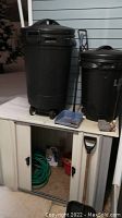 Photo showing metal shed with doors open revealing hose and items. Two black plastic waste cans on top of shed. Blue dustpan with brush and shovel placed next to cans.