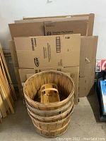 Photo showing multiple flattened corrugated cardboard boxes in various sizes stacked vertically against a wall with four wooden slatted bushel baskets stacked in front.