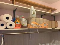 Shelf with full case of Cascades roll towels plus several loose rolls, Xtra brand dish soap, a jug of vinegar, and white jug with liquid