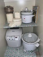 Photo showing all appliances in shelf, including Sunbeam food chopper, Rival slow cooker, Oster bread maker, Westinghouse slow cooker, and slow cooker cookbook.