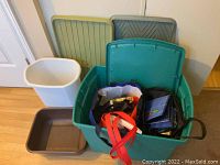 Photo shows green plastic storage tote with hinged lid slightly open containing multiple reusable shopping bags, with two gray plastic boot trays and a white plastic waste can positioned behind and beside it.