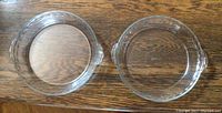 Two clear Pyrex glass pie plates side by side on a wood surface, each with handles and subtle decorative edge detail, showing size and condition