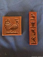 Front view of two beeswax art pieces, a square with a rooster and a rectangular with four designs.