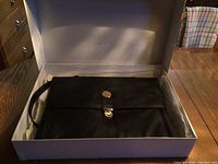 Black leather briefcase in its box showing fold flap with metal lock and handle on the side.