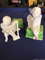 Photo of two cherub figurines on green cushions, one playing harp, one seated blowing kiss, on blue cloth background.