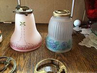 Three vintage glass light fixtures with hand-painted designs and four brass applicators placed on a wood surface.