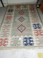 Full view of large wool rug with tribal and diamond shape motifs in muted tones of blue, red, green, and beige on a cream background.