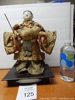 Full front view of the large antique Japanese doll showing its gold patterned kimono and white painted face with hands holding a wooden rod. Water bottle placed next to doll for size reference.