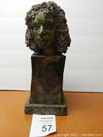 Front view of painted hard clay bust sculpture of a woman with textured curly hair on rectangular pedestal base.