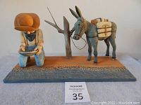 Full view of wood sculpture showing the miner kneeling with pan, the mule with saddlebags, and a tree on a rectangular base.