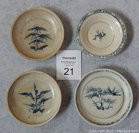 Four small ceramic plates arranged in a square pattern on a fabric surface alongside a MaxSold auction card labeled 21. Each plate displays different blue underglaze designs. Plates show visible signs of age and wear.