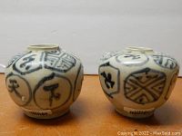Pair of two small porcelain jars side by side showing blue circular character decoration and glossy glaze. Inventory stickers visible on jar bases