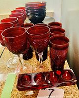 Various red glass drinking glasses and wine glasses with textured cut patterns shown alongside rectangular candle holders with four round candle slots.