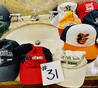 Photo of assorted hats arranged around a sink, showing variety of logos and colors including Baltimore Orioles, Park Tennis New Orleans, conference champion and athletic hats.