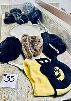 Eight hats arranged on a countertop including black and brown faux fur hats and a yellow and black hat with logo.