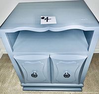 Front view of one light grey-blue wood night stand showing two closed doors with silver tone round knobs and an open shelf above them. The top edge has a scalloped shape.