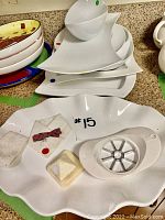 Large white ceramic platter with wavy edges displaying a small chip. Several kitchen accessories including an apple slicer, shirt decoration, small cloth, and soap holder placed on the platter.