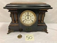 View of the full antique SF mantle clock showing its black body, fluted columns with gold accents, lion head decorations, and a detached pendulum piece in front.