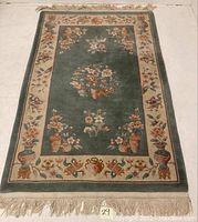 Full view of rectangular wool area rug with green center field and beige floral border, fringed ends.
