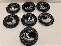 Seven black art glass plates with reticulated edges and hand-painted Mary Gregory style scenes featuring children and dogs in outdoor settings.