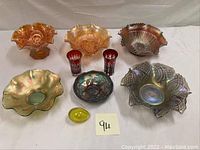 Full lot of carnival glass bowls, ruby red glasses, and yellow bubble glass egg paperweight arranged on white surface.