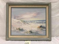 Full view of framed oil painting showing lighthouse on left, beach, dunes, and grasses in muted pastel tones.