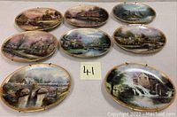 Eight oval Thomas Kinkade collector’s plates arranged on white surface, showing various village, river, barn, and mill scenes all with gold decorative borders.