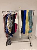 Full view of a clothing rack holding assorted women's clothing on hangers and scarves draped over the rack. Clothing includes various patterns and solid colors. Scarves are in several colors including white, black, blue, yellow, and light blue multicolor patterns.