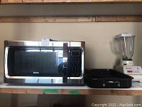 RCA stainless steel microwave next to black roasting pan and Osterizer blender on wooden shelf.