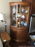 Full view of wooden corner china cabinet with glass upper doors showing displayed china and glassware inside.